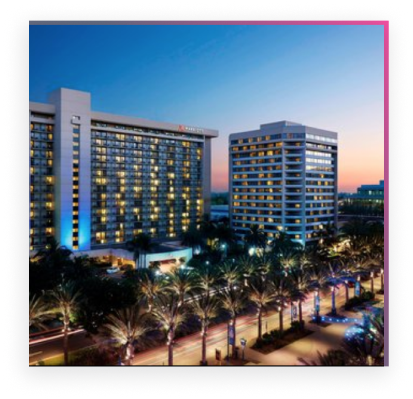 Two modern high-rise hotels are illuminated at dusk, with palm trees lining the busy street in the foreground—an inviting scene for FutureCon OC 2024 attendees.