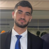 A young man with short dark hair and a trimmed beard is wearing a dark suit, white shirt, and blue tie, standing indoors at a CyberWeek 2024 event.