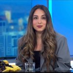 A woman with long, wavy hair sits at a news desk with a microphone, a glass of water, and a plate of fruit in front of her; the Cybertech Tel Aviv 2025 cityscape blurred in the background.